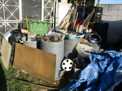 Photo 48: Dismantled machinery parts, discarded board and wood materials, tarps, and bags of trash are maintained on a residential property.
